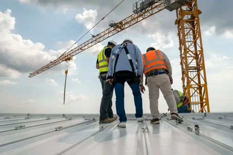 Three construction workers in safety vests standing on a metal rooftop near a crane.